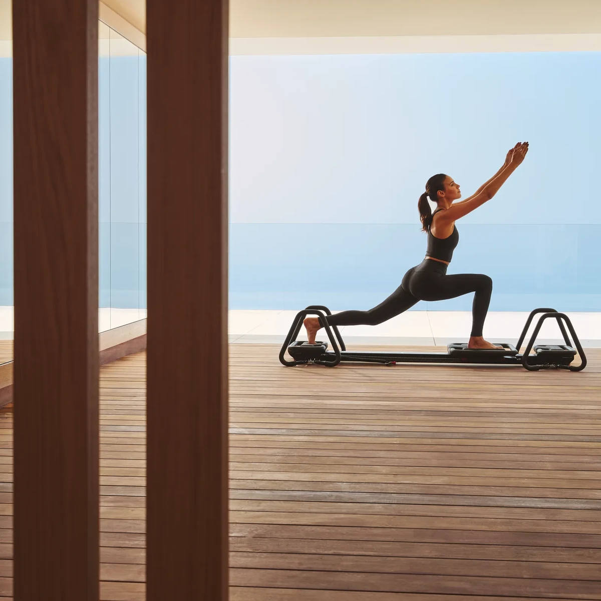 Guest performing a lunge on a Pilates reformer in a serene wooden studio overlooking the sea.