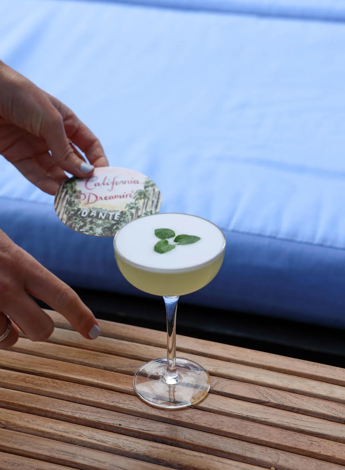 Hand holding “California Dreamin’” coaster above frothy cocktail garnished with mint leaves.