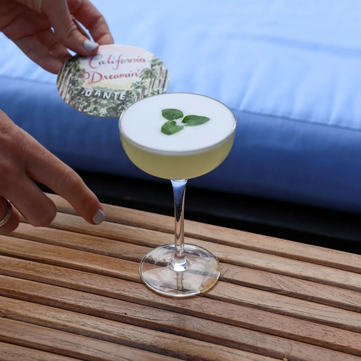Hand holding “California Dreamin’” coaster above frothy cocktail garnished with mint leaves.