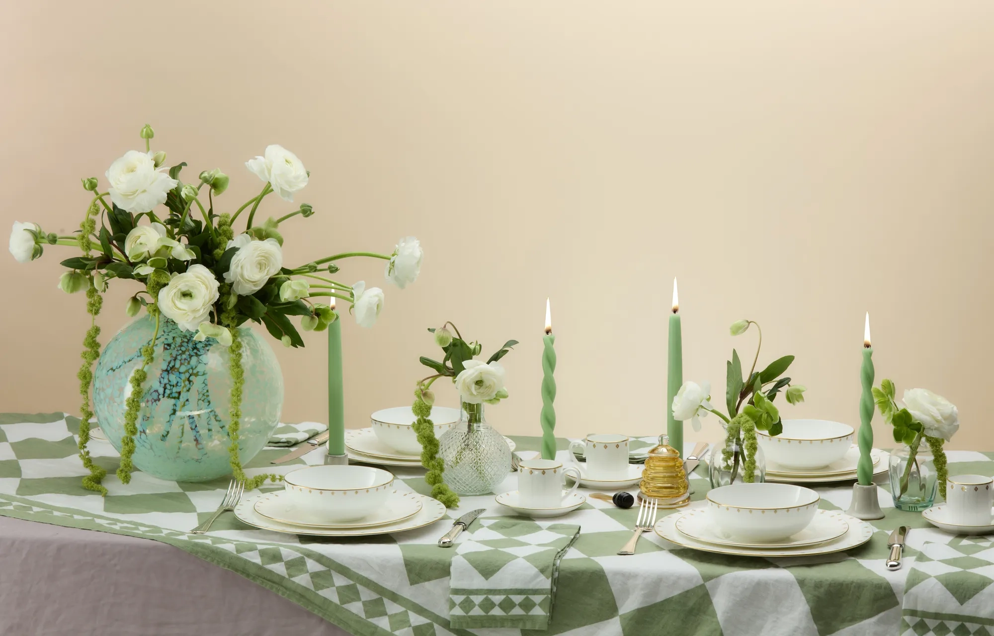 A beautifully arranged dining table set with fine white and gold-rimmed china, green patterned linens, pastel green twisted candles, and vases of white ranunculus flowers in soft, natural lighting.