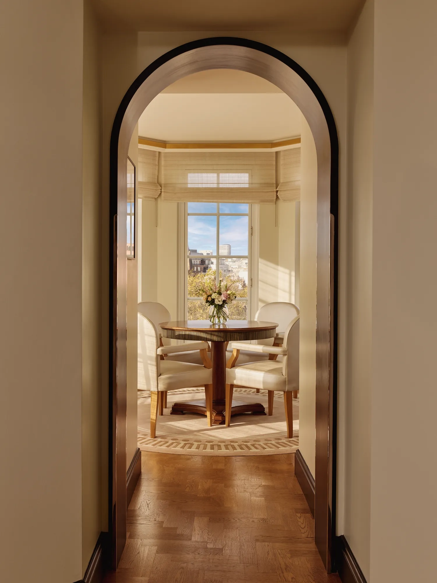 View through an arched doorway leading to a bright dining alcove with four cream chairs around a round wooden table and large windows overlooking the city.