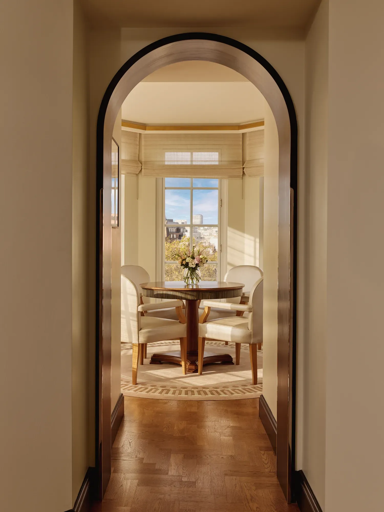 View through an arched doorway leading to a bright dining alcove with four cream chairs around a round wooden table and large windows overlooking the city.