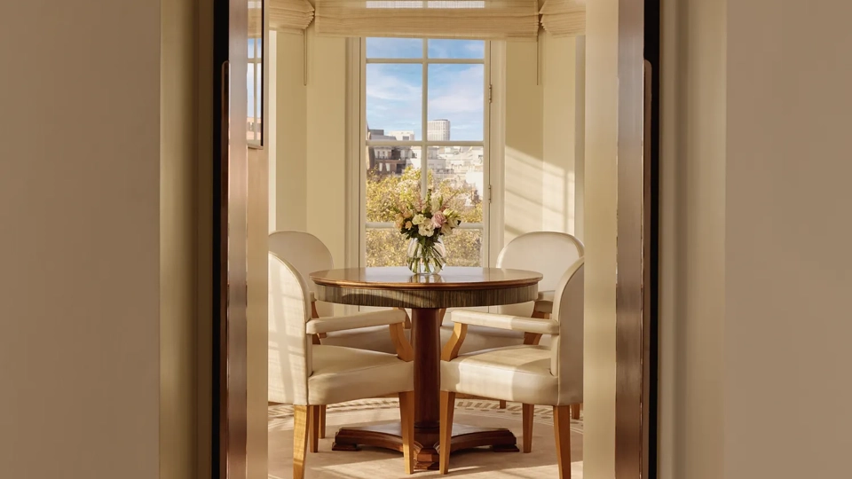 View through an arched doorway leading to a bright dining alcove with four cream chairs around a round wooden table and large windows overlooking the city.