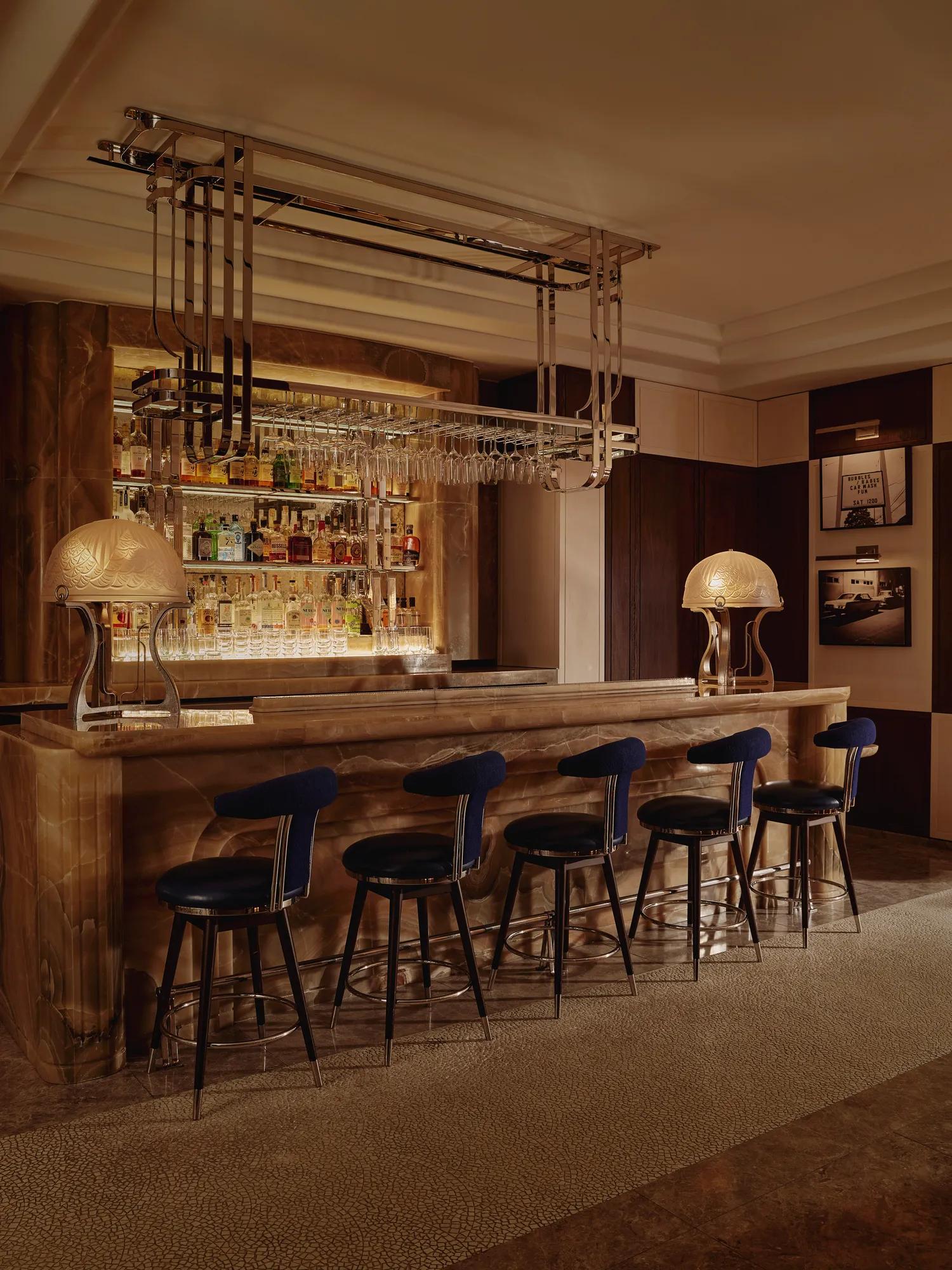 Onyx bar with chrome glass rack, two sculptural lamps and five navy barstools in a dim lounge.