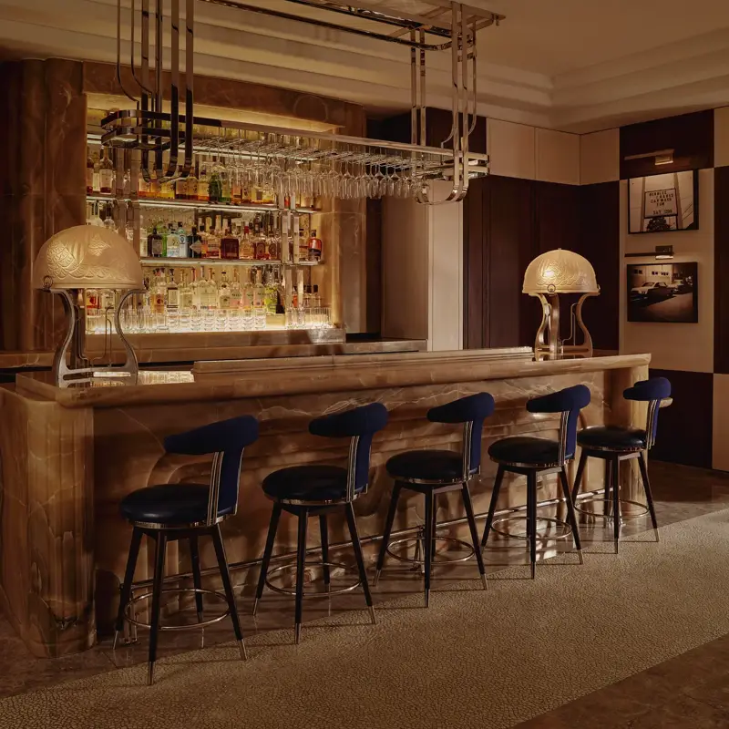 Onyx bar with chrome glass rack, two sculptural lamps and five navy barstools in a dim lounge.