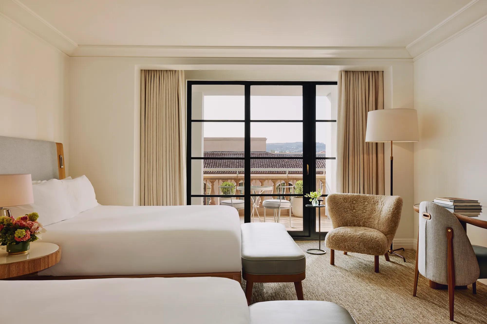 Two-queen room facing a balcony, with benches, lounge chair, floor lamp, and writing desk near sliding doors.