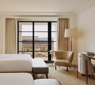 Two-queen room facing a balcony, with benches, lounge chair, floor lamp, and writing desk near sliding doors.