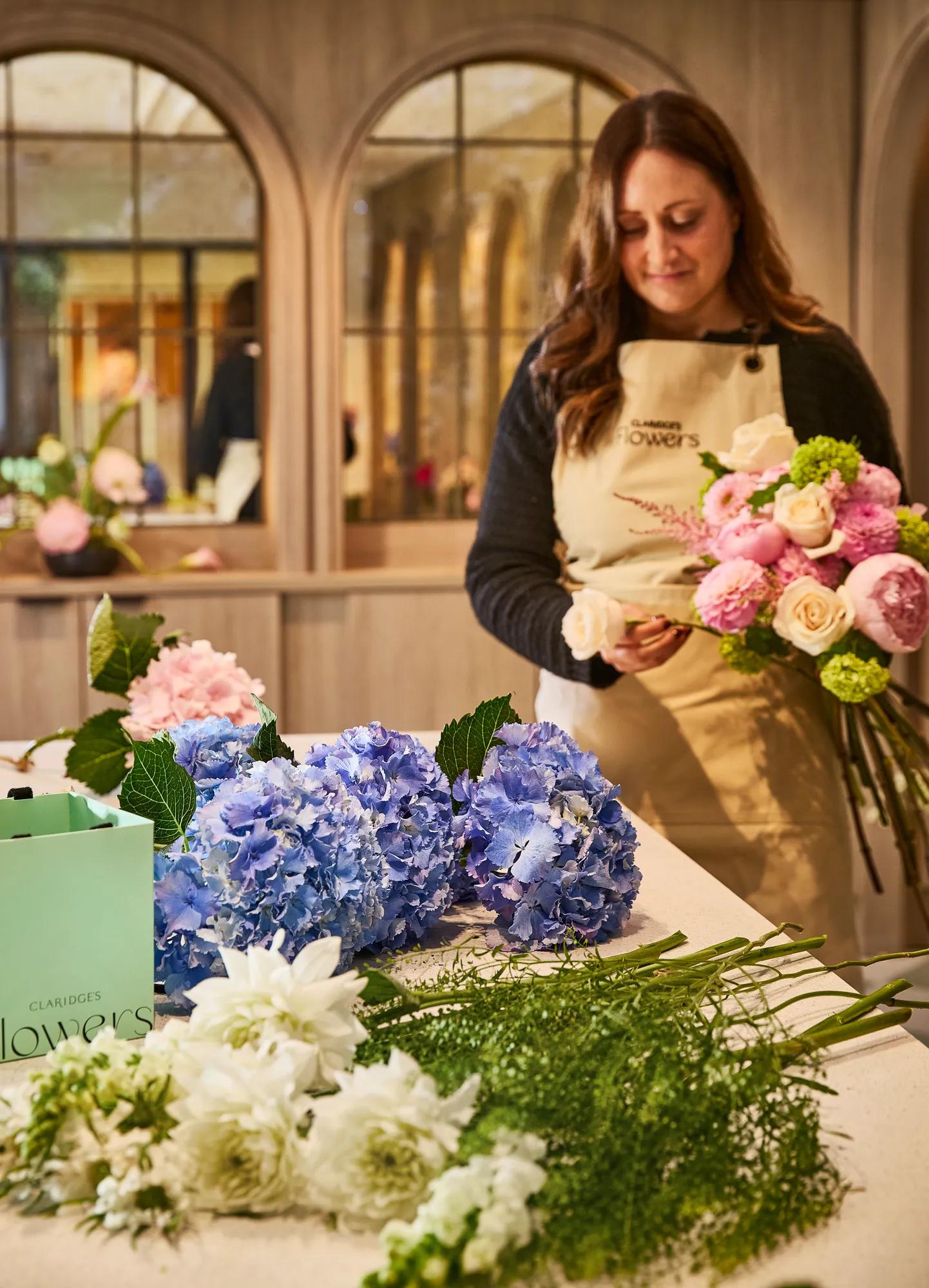 **Alt text (WCAG 2.2 AA, under 125 characters):**

Florist arranging a bouquet beside blue hydrangeas and seasonal blooms on a worktable inside a warm, wood-panelled boutique.
