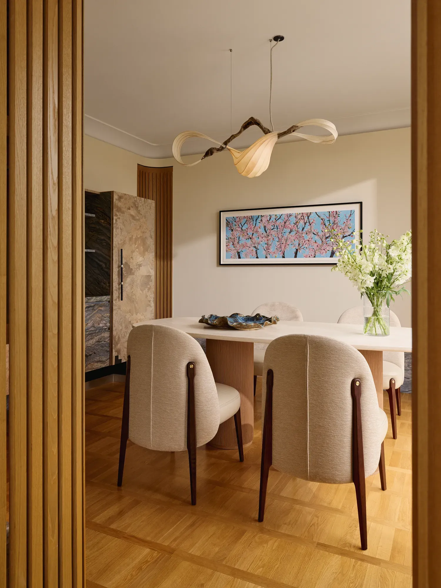 Elegant dining space with a white marble table, upholstered chairs, and a sculptural chandelier above.