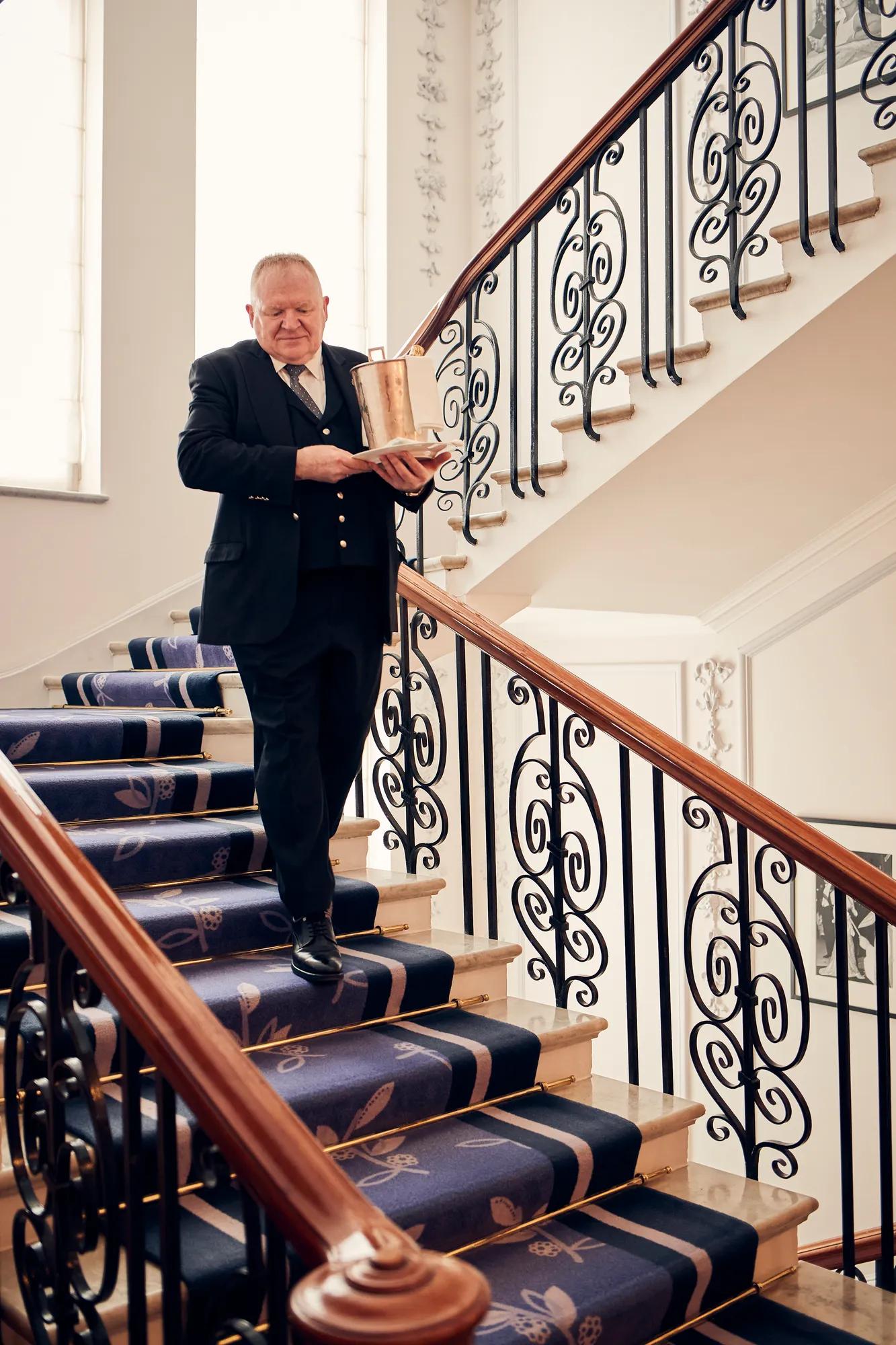 A hotel staff member in a formal navy uniform walks down a grand staircase with decorative wrought-iron railings and a patterned blue carpet, carrying a gold ice bucket with a bottle of champagne.