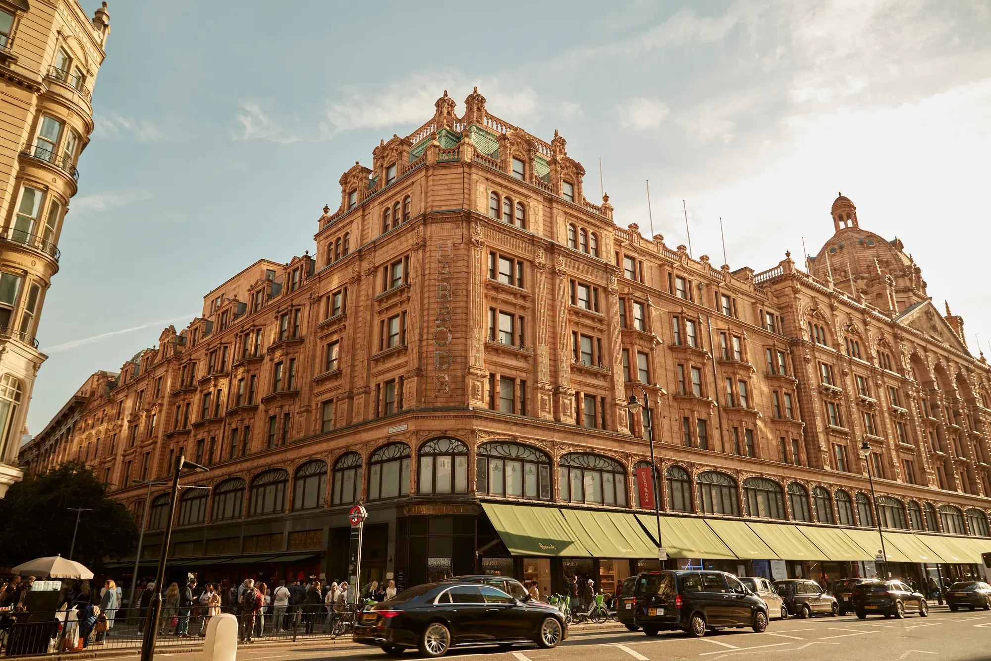 Harrods exterior with ornate terracotta facade, green awnings, and busy street with cars and shoppers.