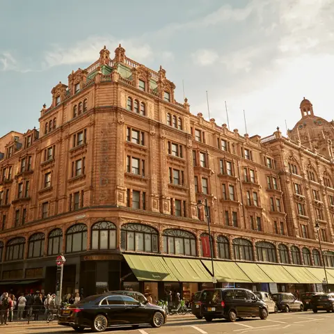 Harrods exterior with ornate terracotta facade, green awnings, and busy street with cars and shoppers.