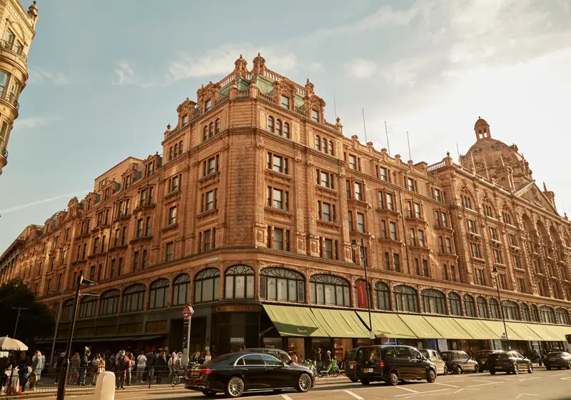 Harrods exterior with ornate terracotta facade, green awnings, and busy street with cars and shoppers.