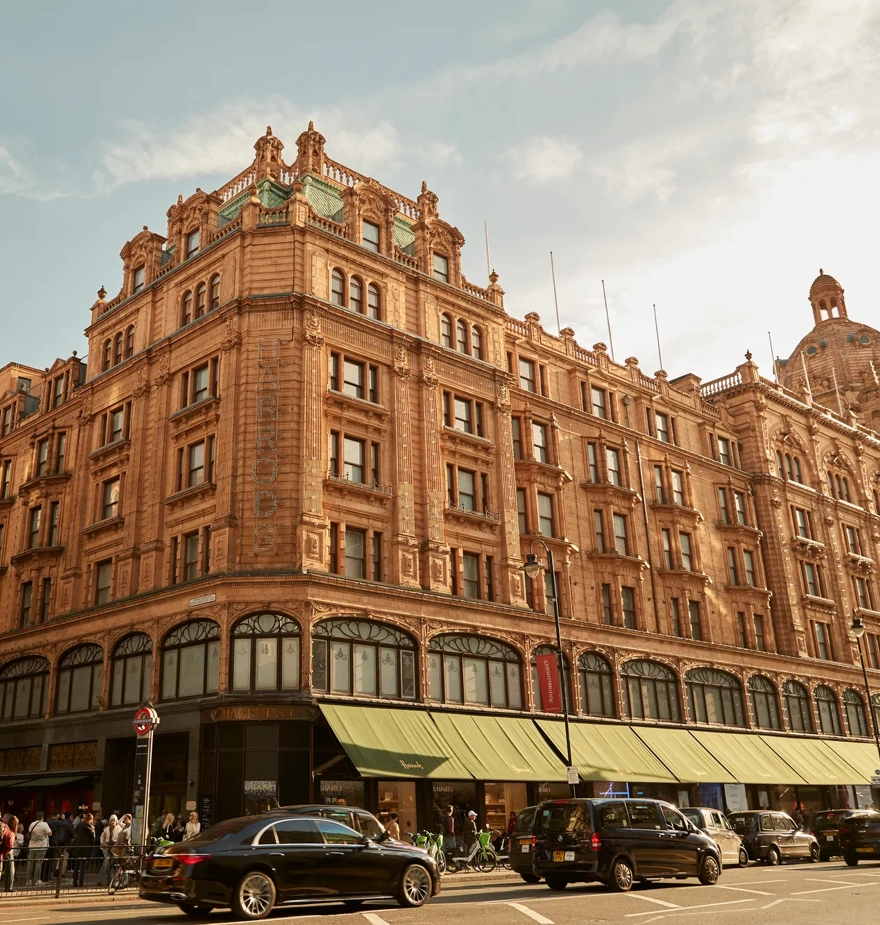 Harrods exterior with ornate terracotta facade, green awnings, and busy street with cars and shoppers.