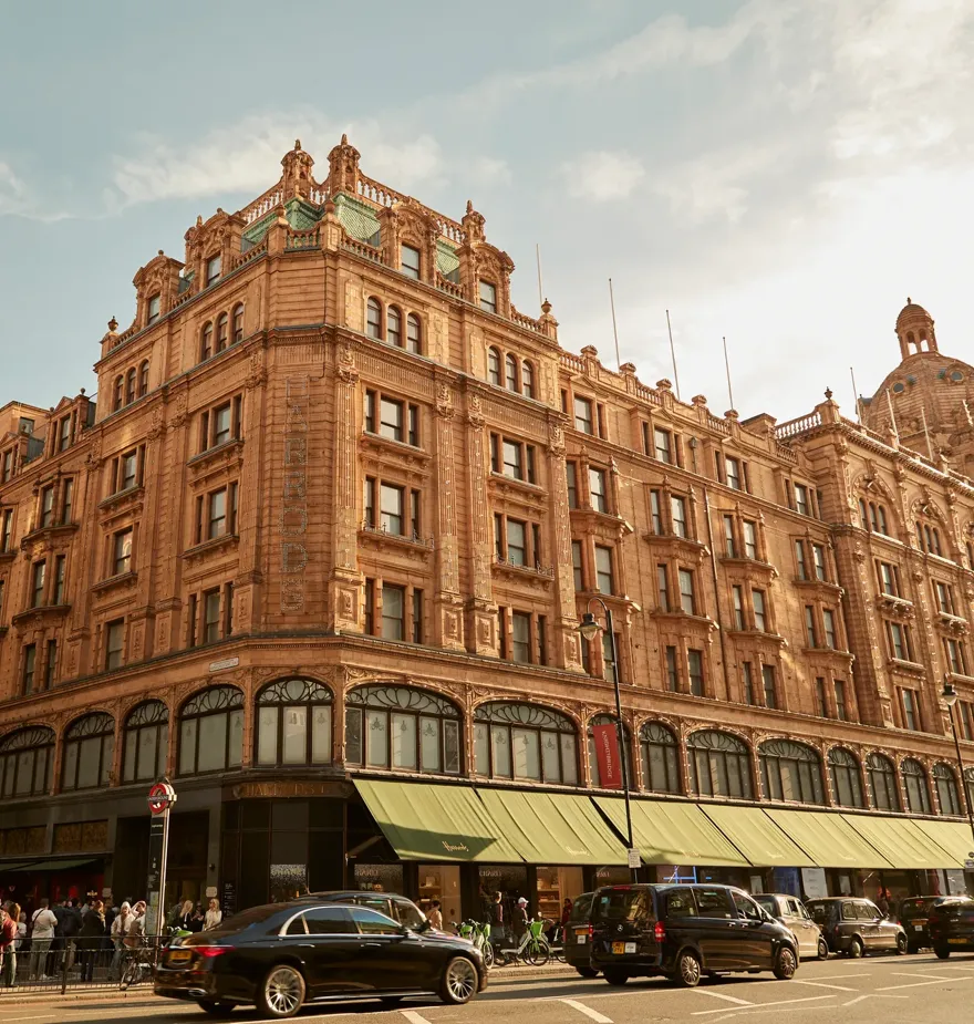 Harrods exterior with ornate terracotta facade, green awnings, and busy street with cars and shoppers.