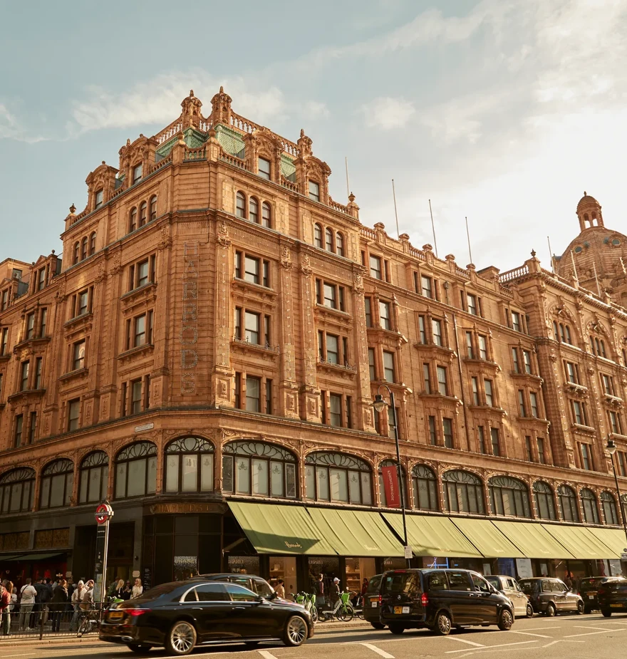 Harrods exterior with ornate terracotta facade, green awnings, and busy street with cars and shoppers.