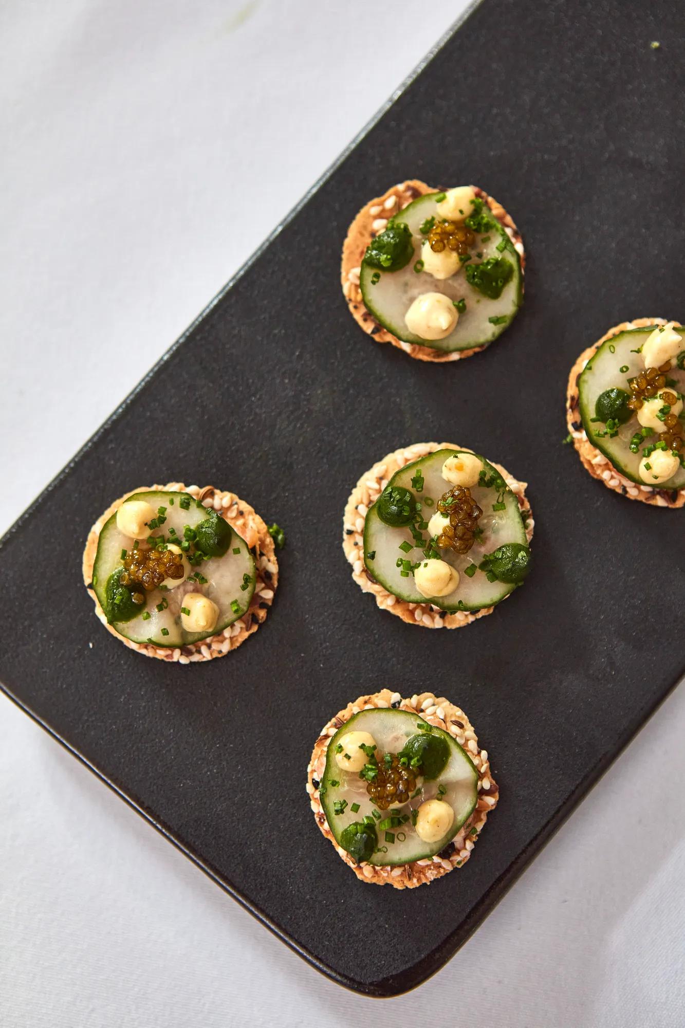 Close-up of canapés topped with cucumber, herbs, cream, and caviar on a black platter.