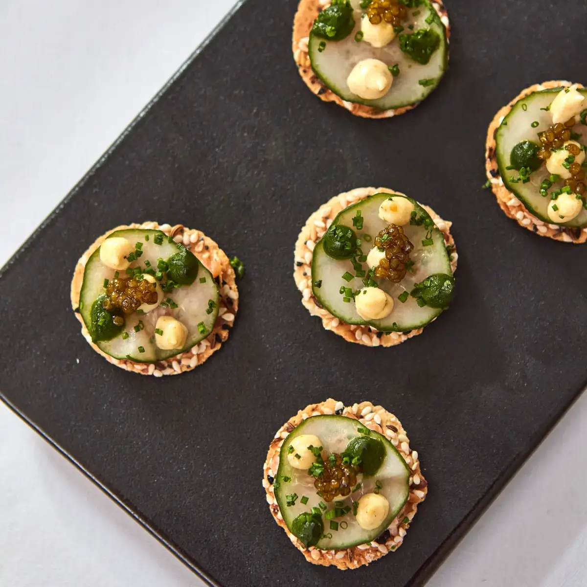 Close-up of canapés topped with cucumber, herbs, cream, and caviar on a black platter.