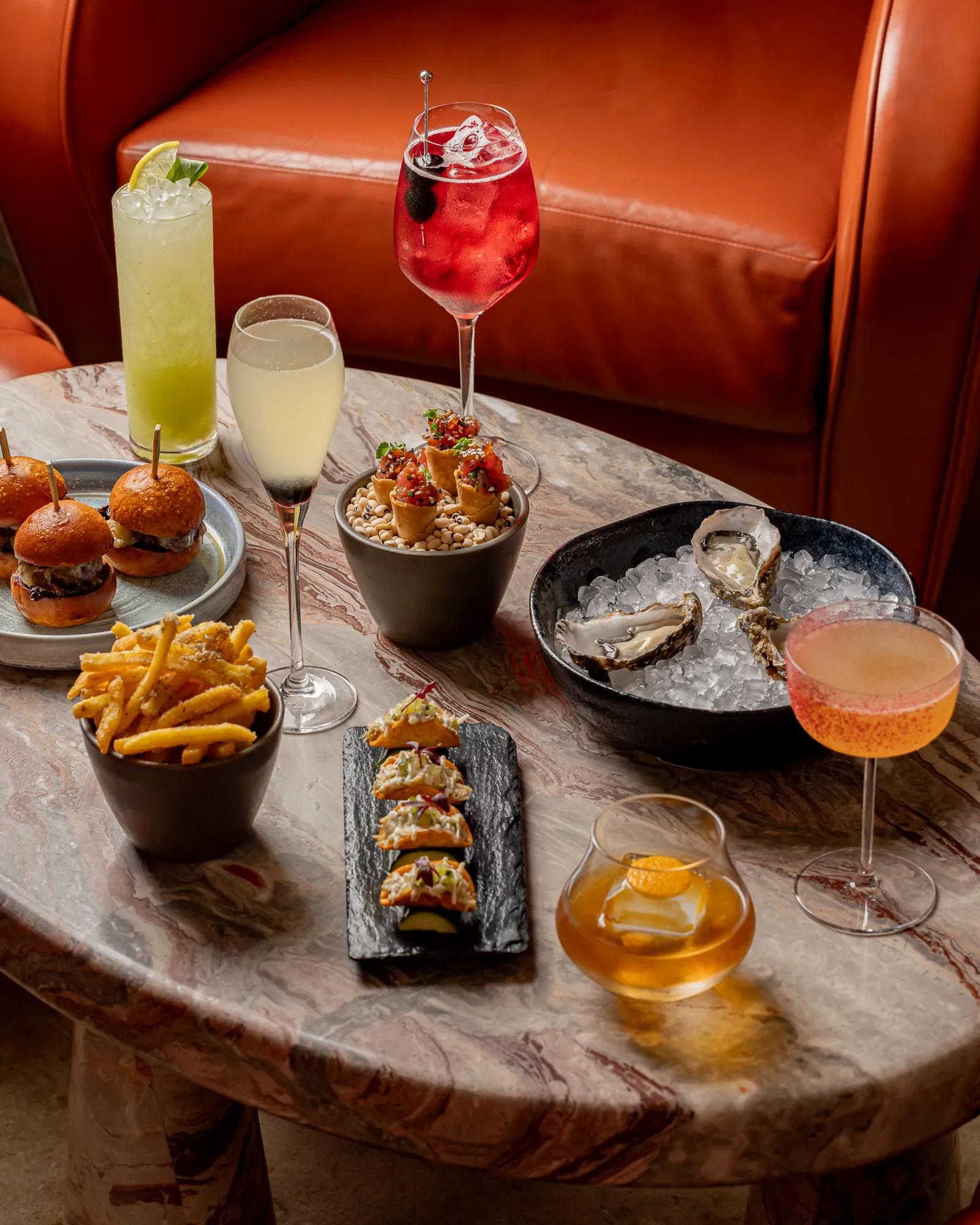 Marble table with oysters, sliders, fries, small tacos, and assorted cocktails in colourful glassware.