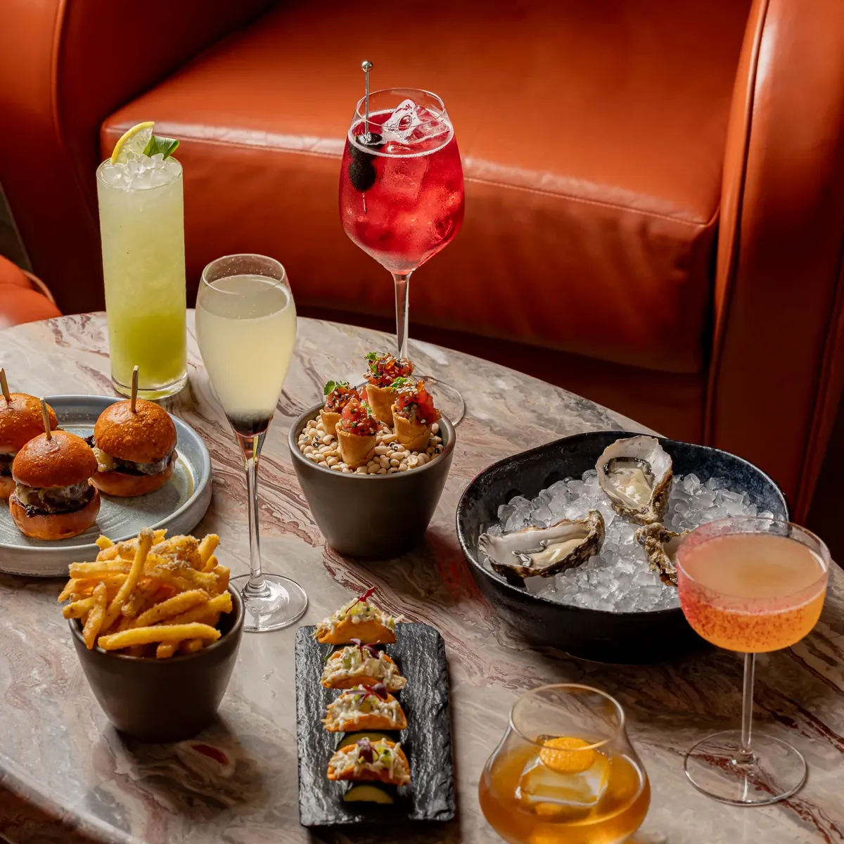 Marble table with oysters, sliders, fries, small tacos, and assorted cocktails in colourful glassware.