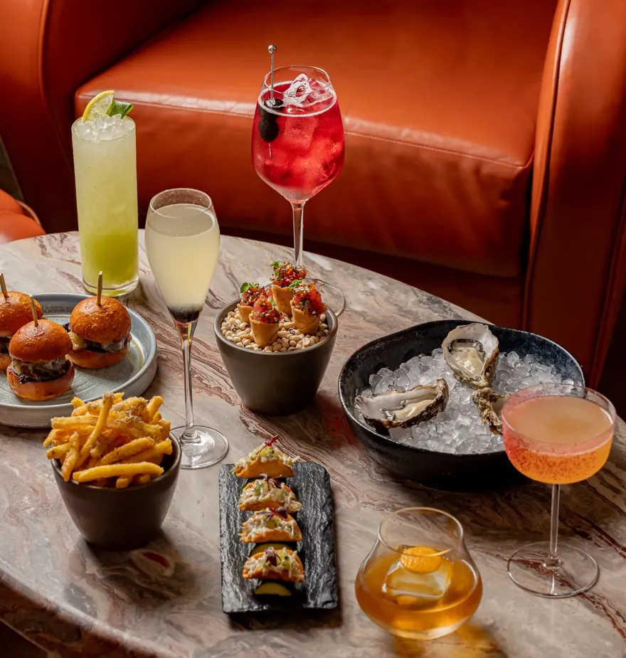 Marble table with oysters, sliders, fries, small tacos, and assorted cocktails in colourful glassware.
