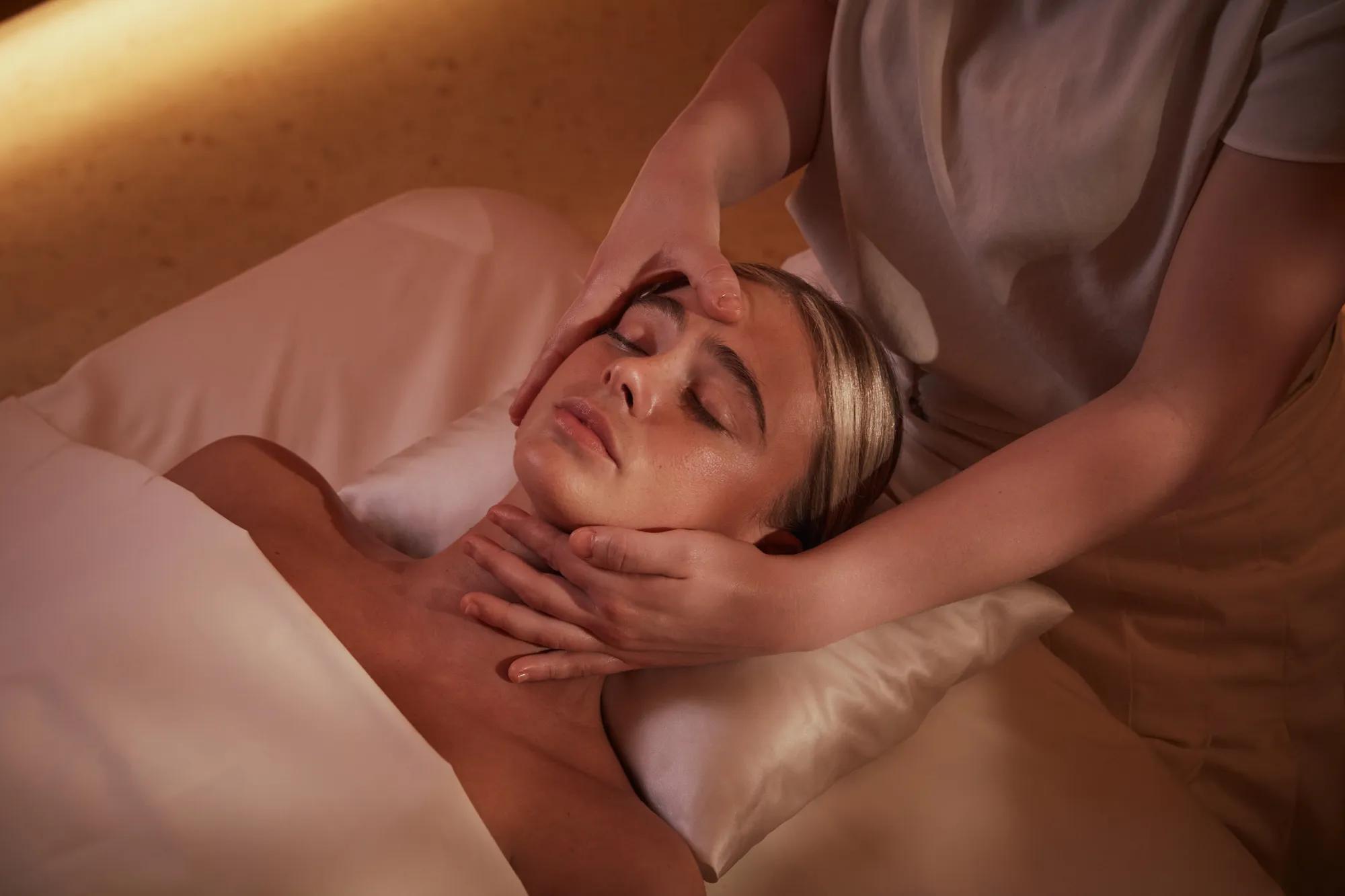 Guest receiving facial massage at spa, lying on pillow under soft warm lighting.