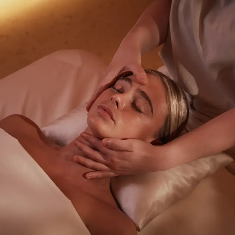 Guest receiving facial massage at spa, lying on pillow under soft warm lighting.