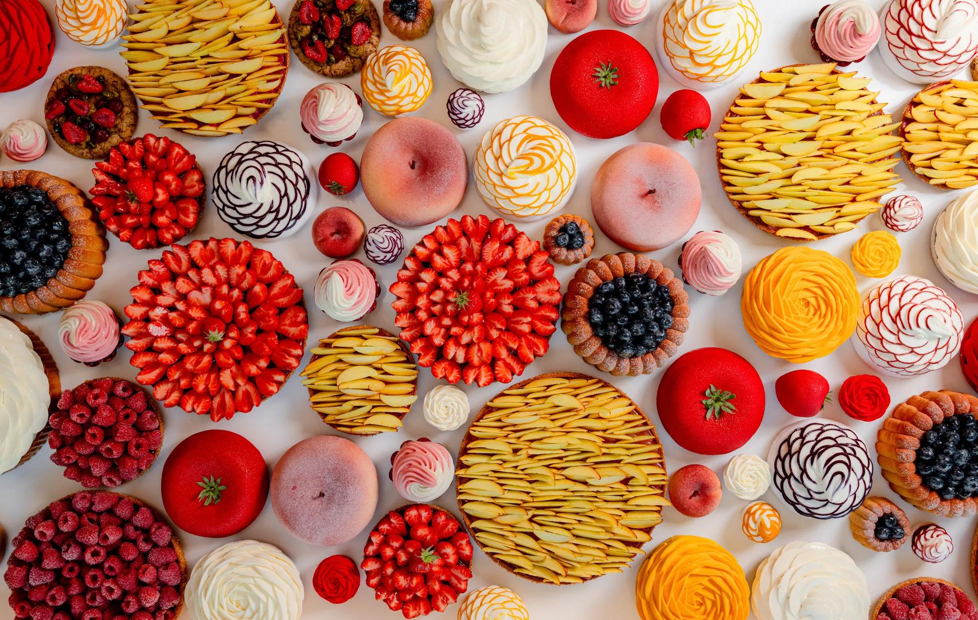Decorative arrangement of fruit tarts, meringues and pastries viewed from above, featuring strawberries, raspberries, blueberries, peaches and apples on a light background.