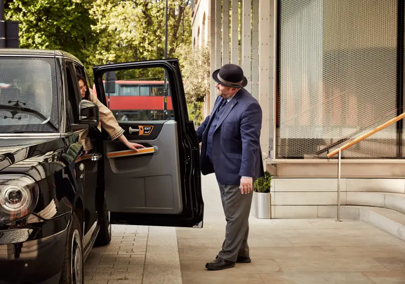 A hotel doorman in a bowler hat opens the door of a black taxi as a guest prepares to step out on the pavement outside the hotel entrance.