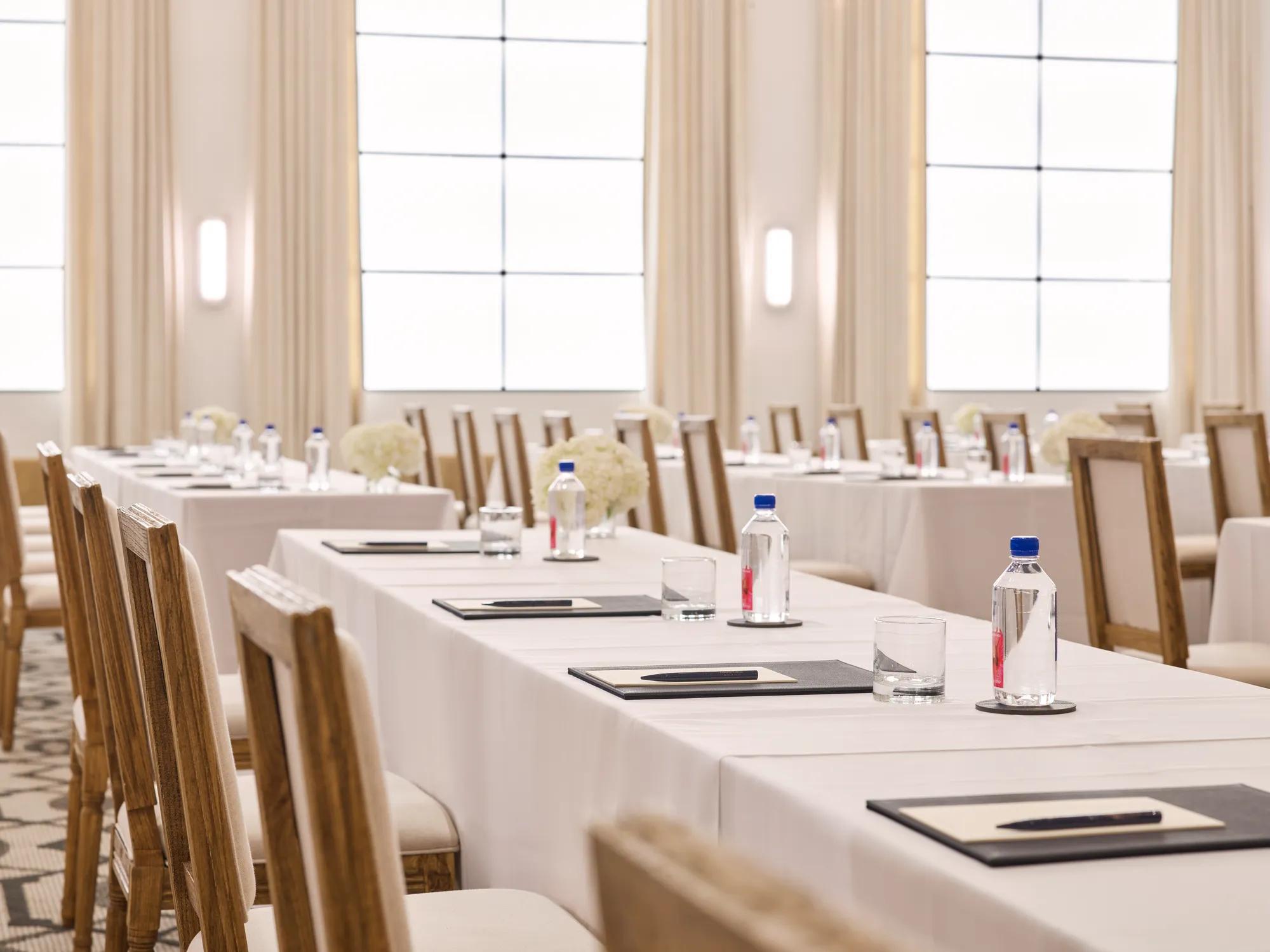 Rows of conference tables with chairs, notepads, and water; bright ballroom with tall drapes and grid windows.