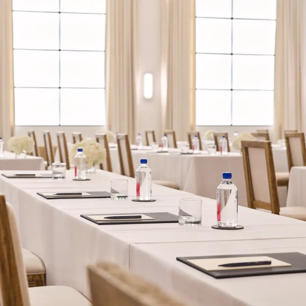 Rows of conference tables with chairs, notepads, and water; bright ballroom with tall drapes and grid windows.