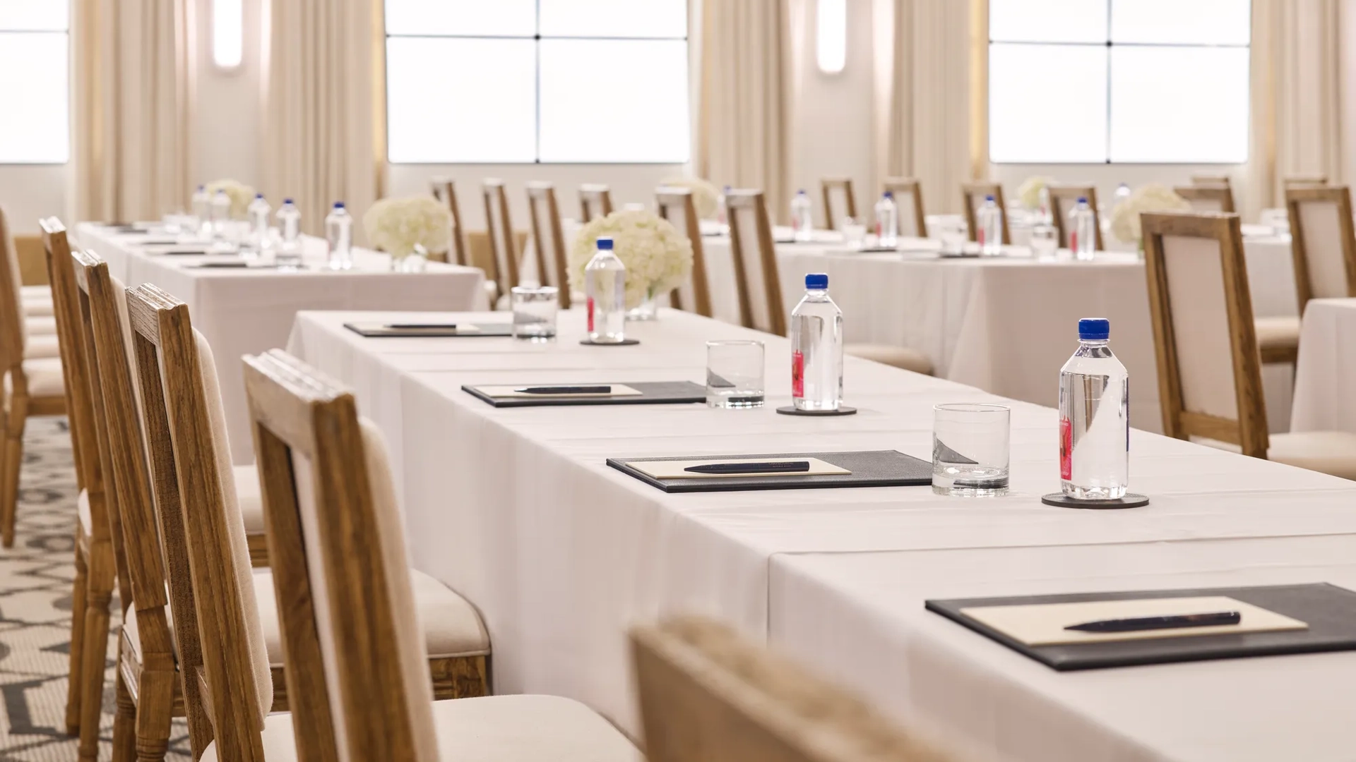Rows of conference tables with chairs, notepads, and water; bright ballroom with tall drapes and grid windows.
