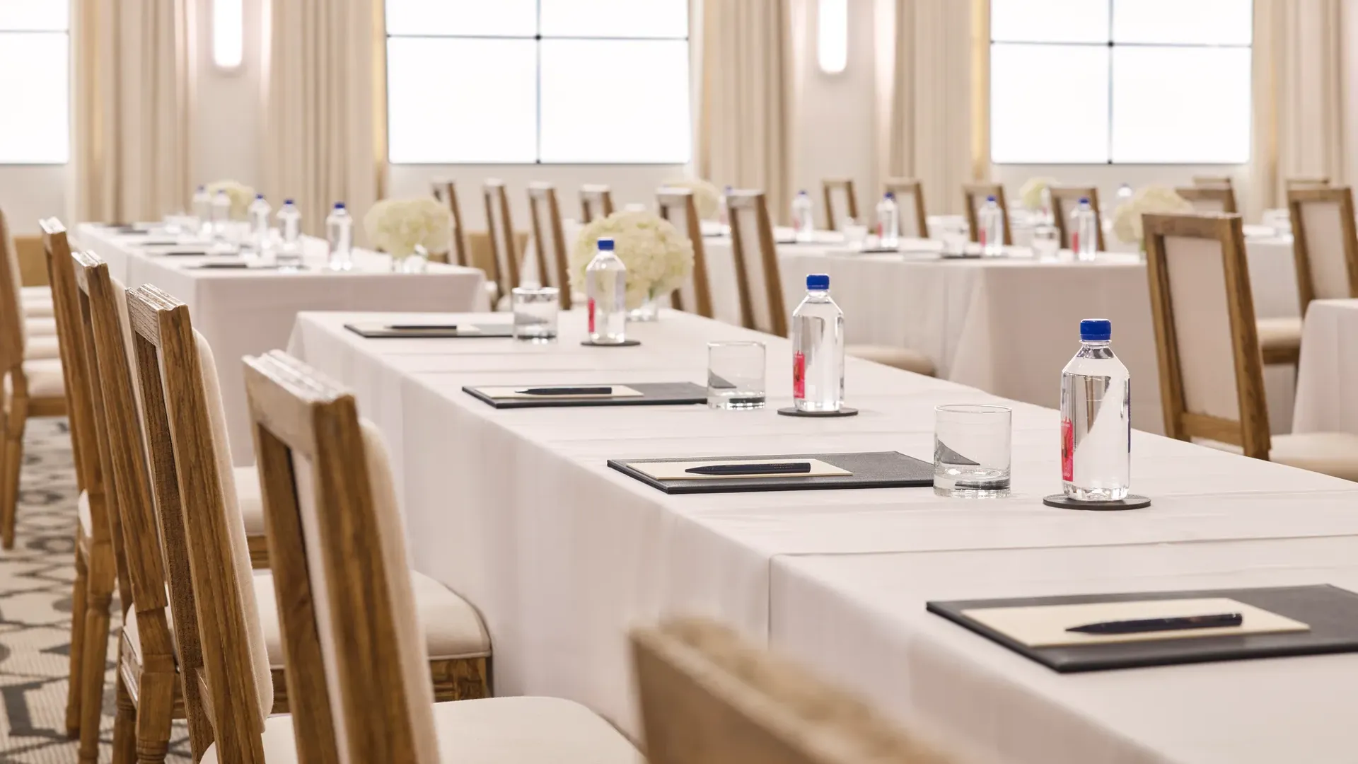 Rows of conference tables with chairs, notepads, and water; bright ballroom with tall drapes and grid windows.