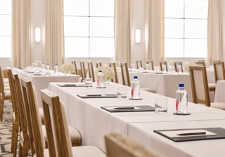 Rows of conference tables with chairs, notepads, and water; bright ballroom with tall drapes and grid windows.