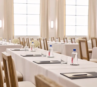 Rows of conference tables with chairs, notepads, and water; bright ballroom with tall drapes and grid windows.