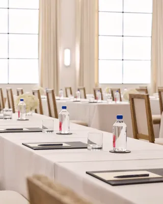Rows of conference tables with chairs, notepads, and water; bright ballroom with tall drapes and grid windows.