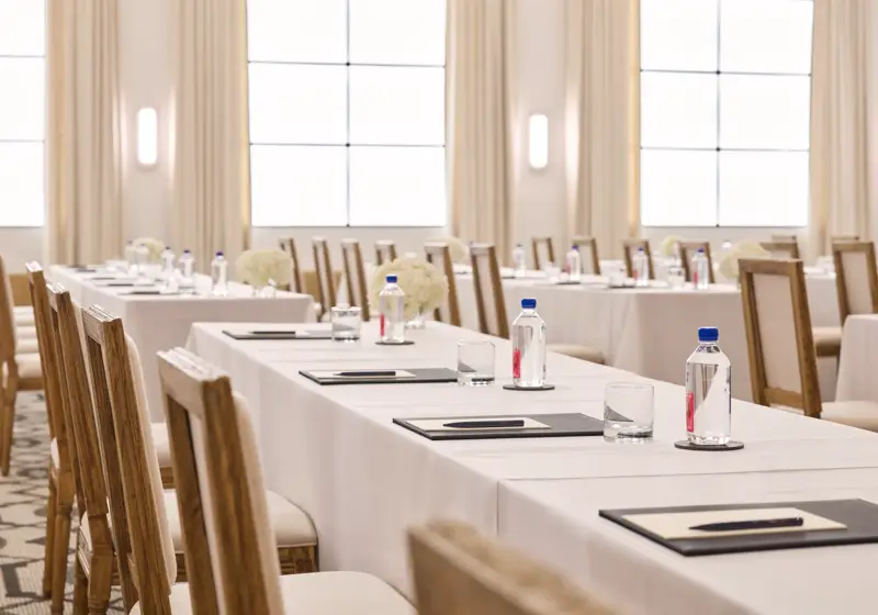 Rows of conference tables with chairs, notepads, and water; bright ballroom with tall drapes and grid windows.