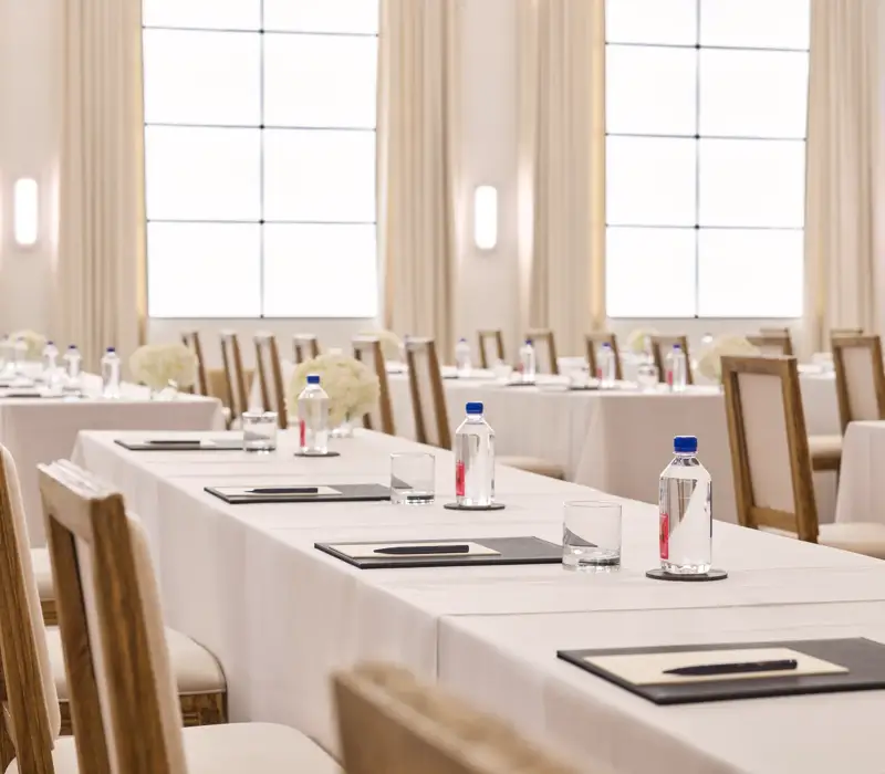 Rows of conference tables with chairs, notepads, and water; bright ballroom with tall drapes and grid windows.