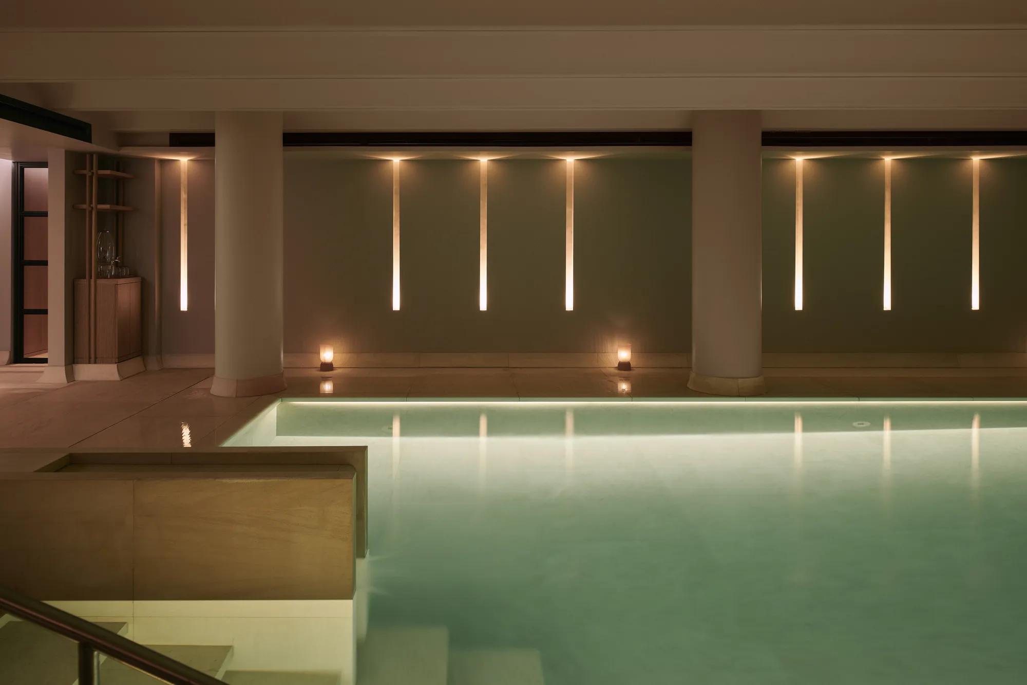Heated indoor swimming pool with pale stone columns, soft lighting and calm, reflective water in a serene spa setting.