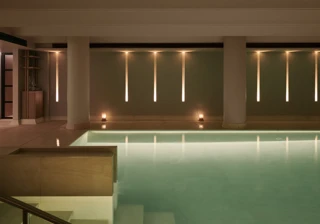 Heated indoor swimming pool with pale stone columns, soft lighting and calm, reflective water in a serene spa setting.