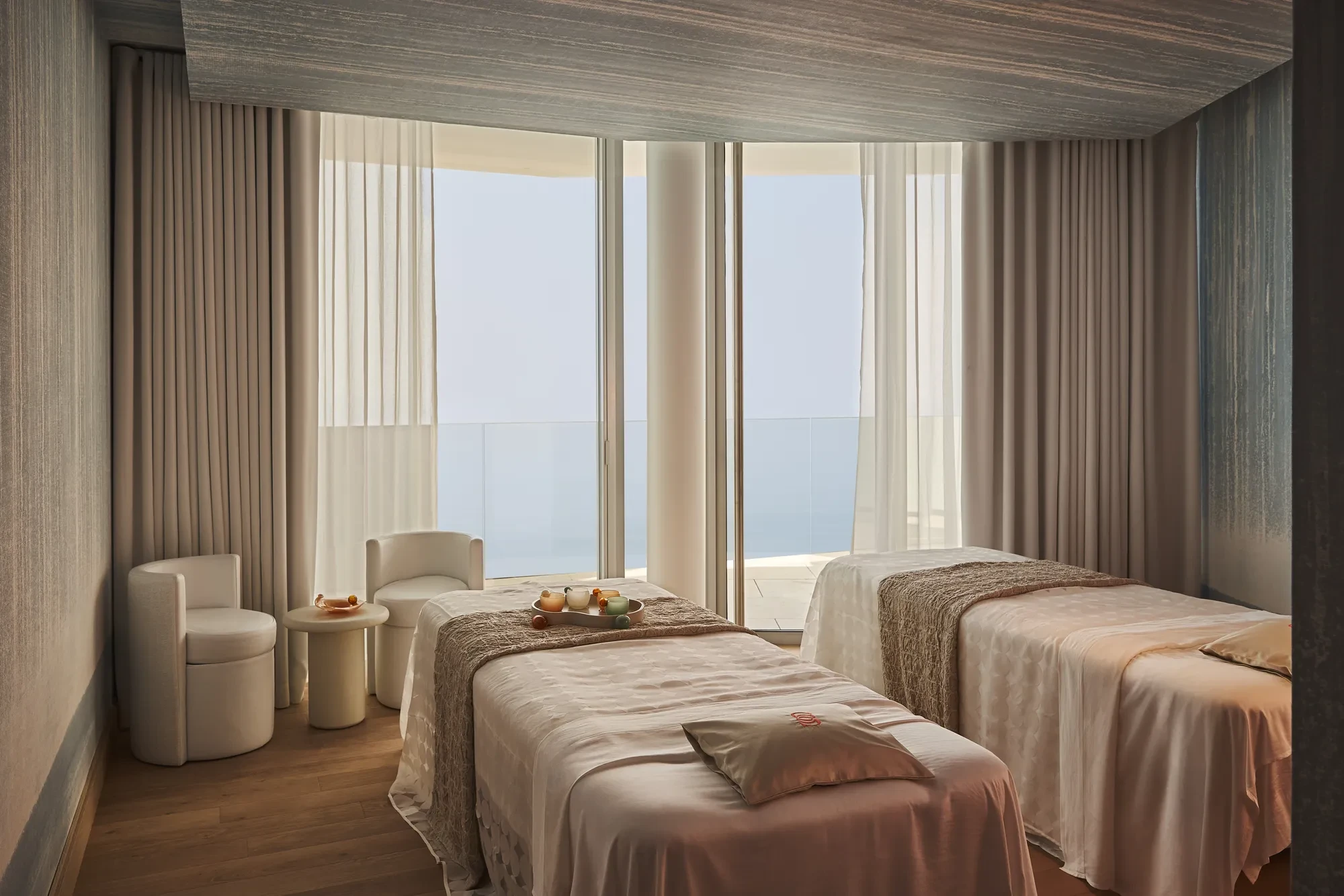 Serene spa treatment room with twin beds, soft textiles, and floor-to-ceiling windows overlooking the sea.
