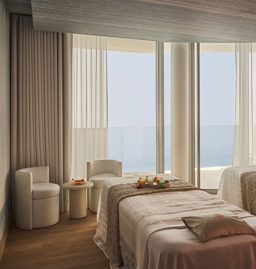 Serene spa treatment room with twin beds, soft textiles, and floor-to-ceiling windows overlooking the sea.