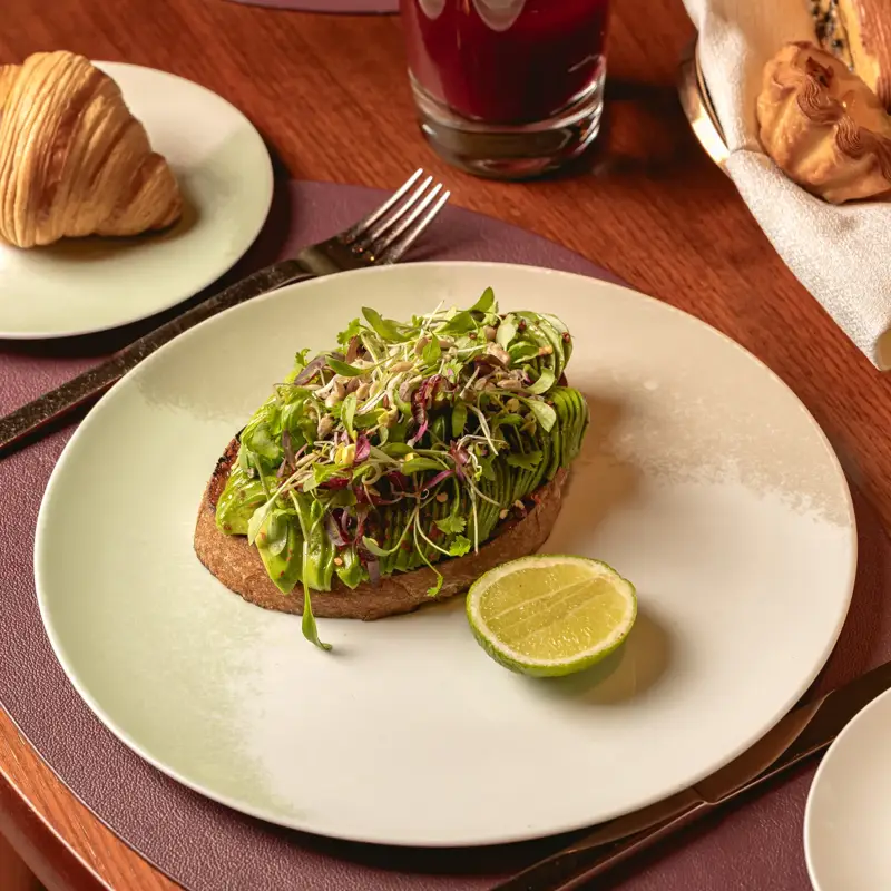 A breakfast plate with avocado toast topped with microgreens and lime, served with pastries and red juice on a wooden table.