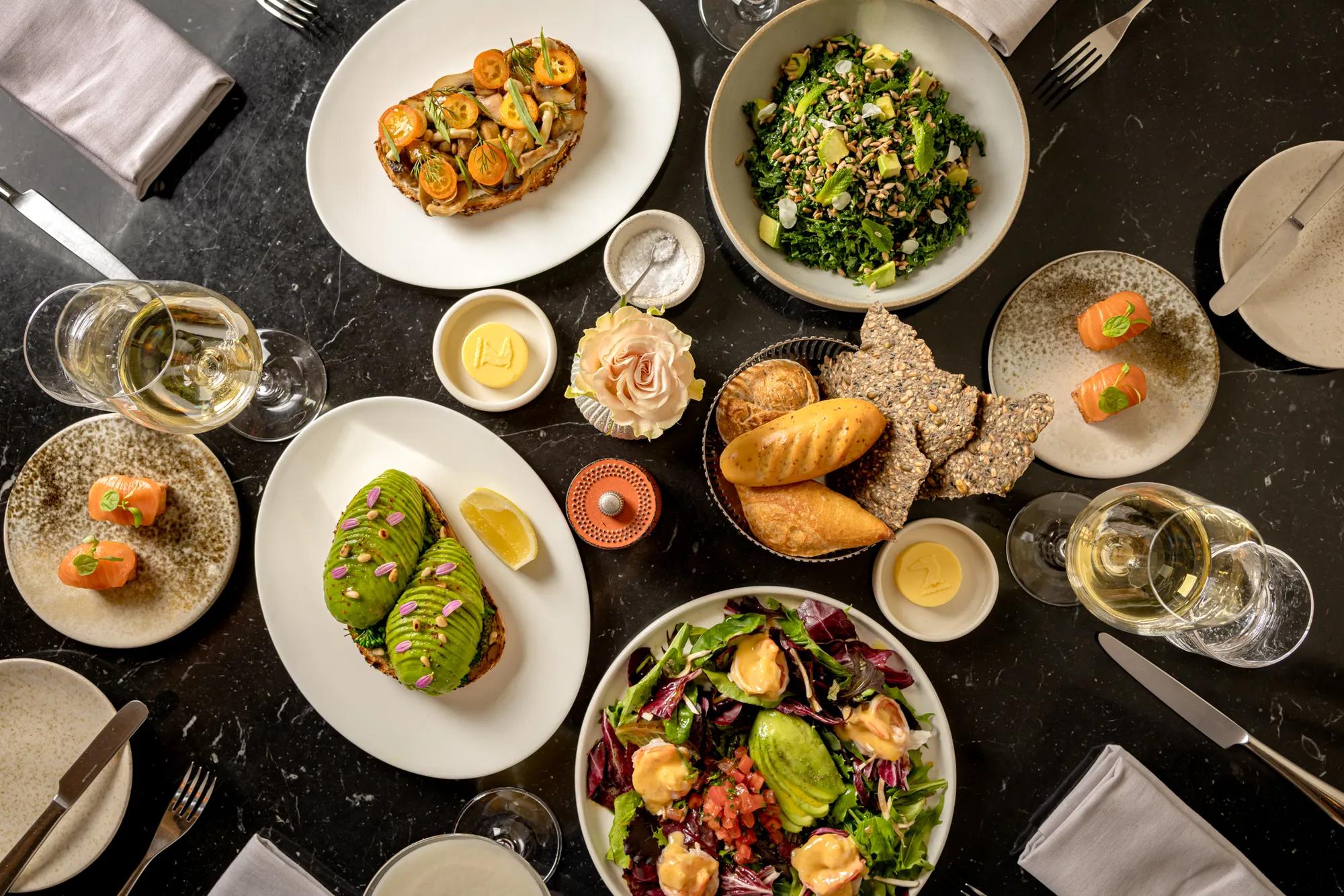 Overhead view of a dining table with salads, bread, avocado toast, seafood appetisers, and wine glasses.