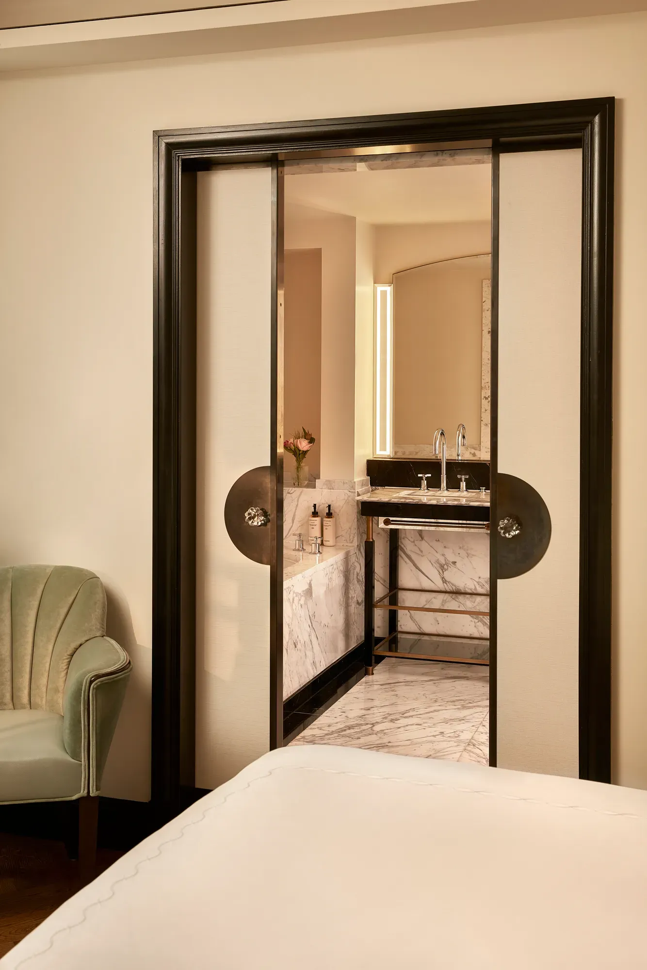 View from the bedroom into a marble bathroom through sliding double doors with dark frames, showing a vanity with twin sinks, a lighted mirror, and a vase of flowers.
