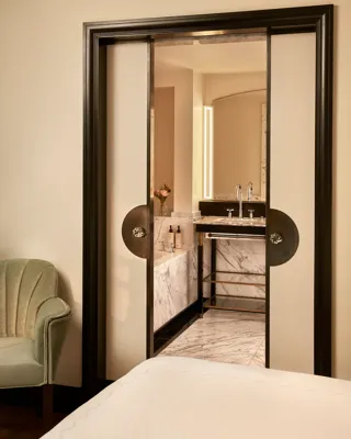 View from the bedroom into a marble bathroom through sliding double doors with dark frames, showing a vanity with twin sinks, a lighted mirror, and a vase of flowers.