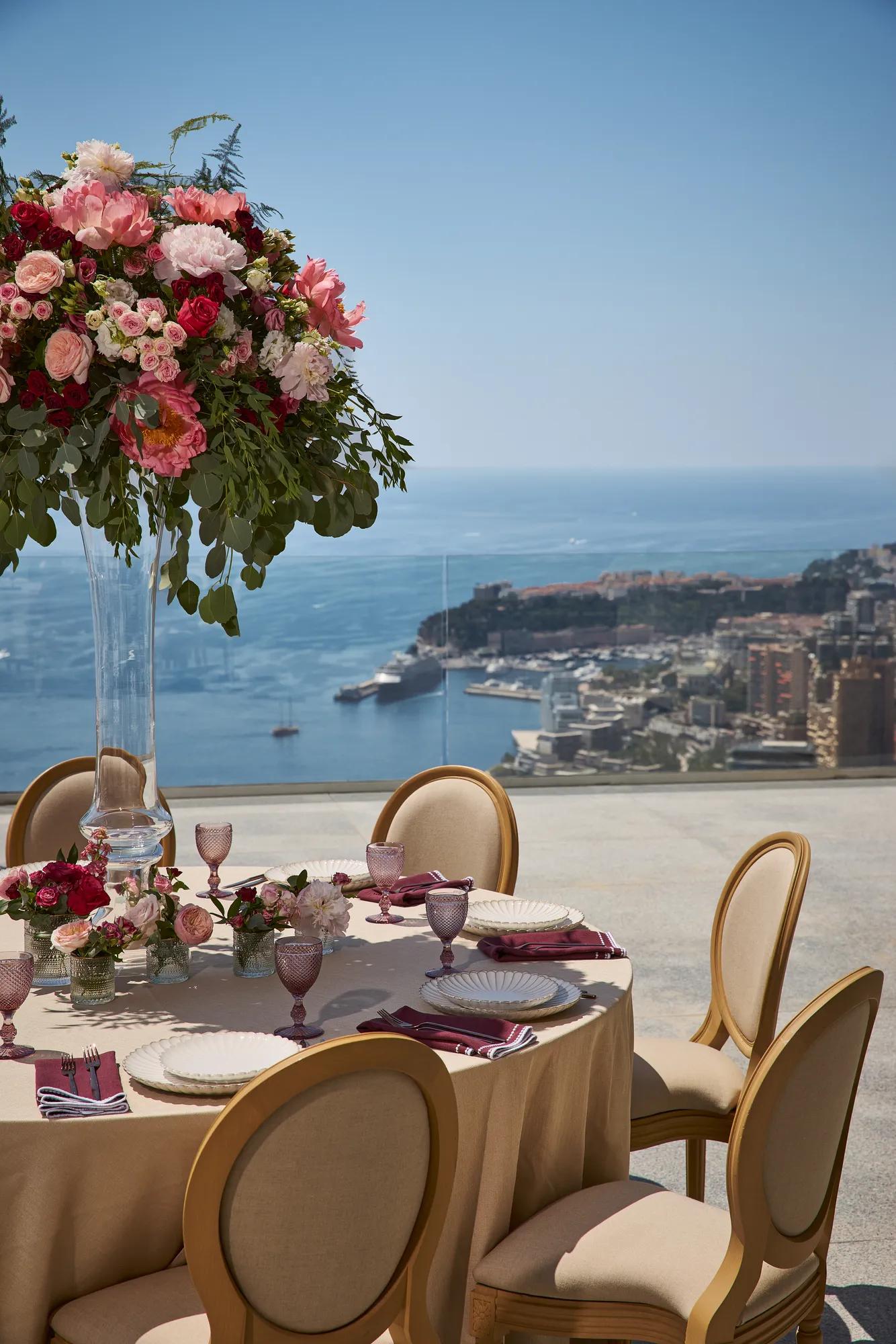 Table ronde en extérieur recouverte d’une nappe beige, entourée de chaises élégantes et de verres roses, ornée d’un grand centre de table floral aux fleurs rouges et roses, avec vue sur la mer et la ville en contrebas.