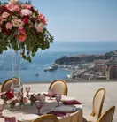 Table ronde en extérieur recouverte d’une nappe beige, entourée de chaises élégantes et de verres roses, ornée d’un grand centre de table floral aux fleurs rouges et roses, avec vue sur la mer et la ville en contrebas.