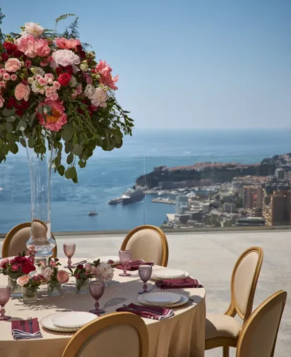 Table ronde en extérieur recouverte d’une nappe beige, entourée de chaises élégantes et de verres roses, ornée d’un grand centre de table floral aux fleurs rouges et roses, avec vue sur la mer et la ville en contrebas.
