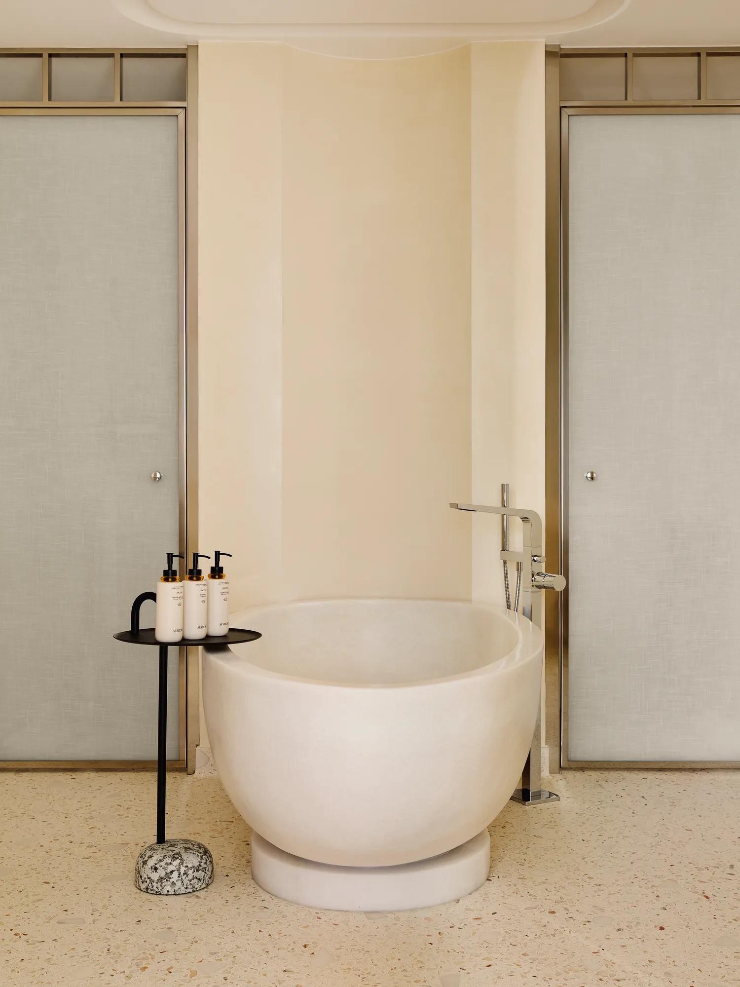 Salle de bain moderne avec baignoire ovale autoportante, robinetterie chromée élégante et petite table avec produits de toilette de luxe.